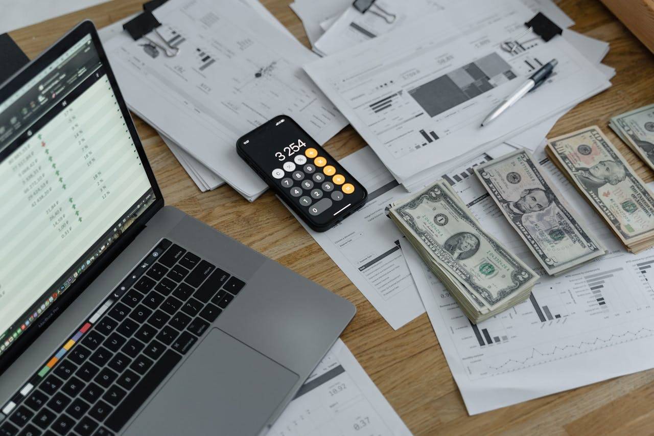 A cluttered desk features a laptop, a smartphone displaying a calculator app, various financial documents, a pen, and several stacks of US dollar bills, all indicating an attempt to organize finances despite having no business income.