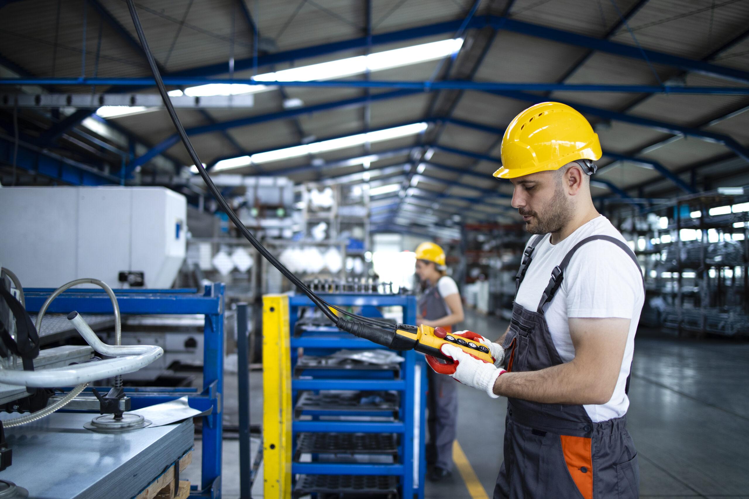 Factory worker wearing uniform and hardhat operating industrial machine with push button joystick in production hall.