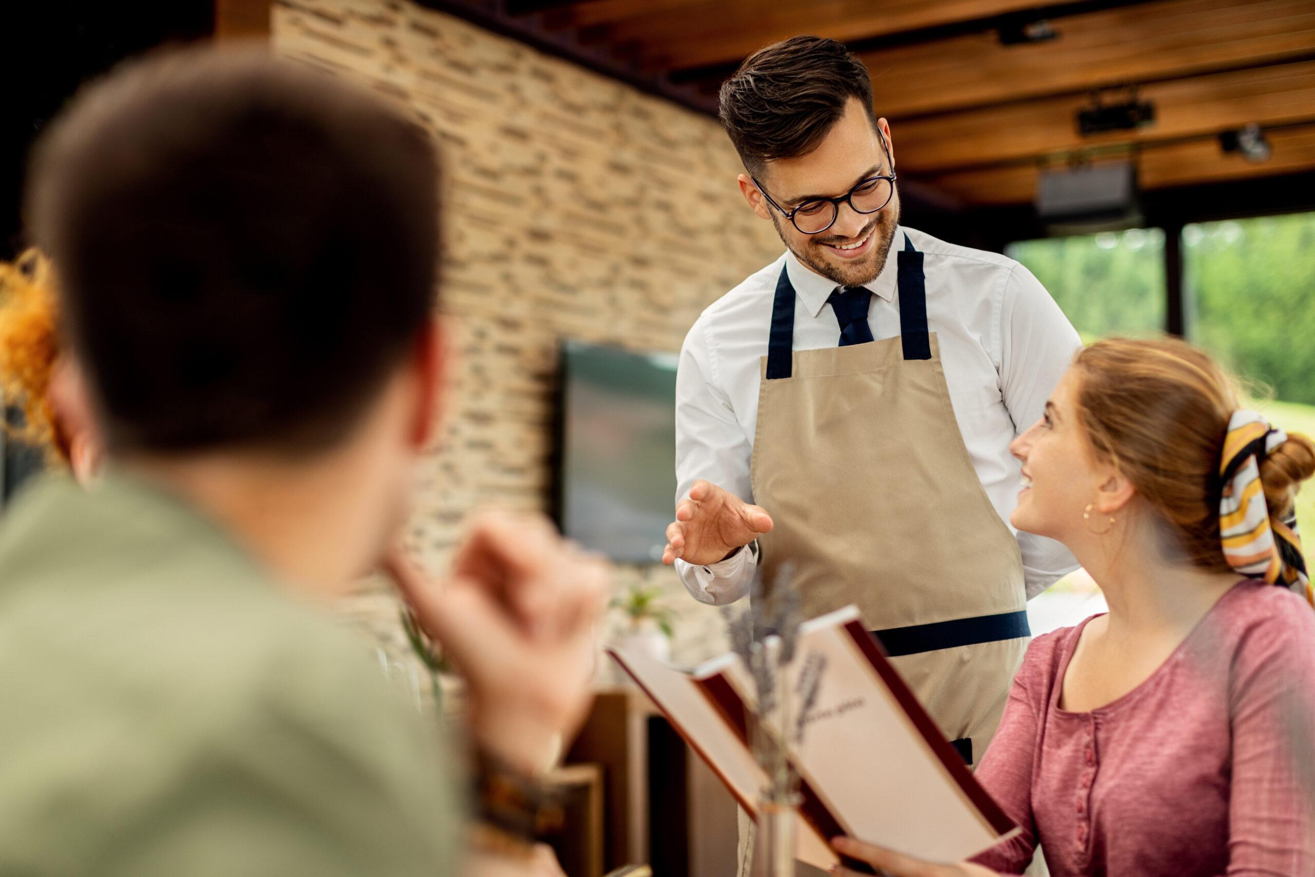 Happy waiter talking to a woman and recommending her what to order from the menu in a restaurant.