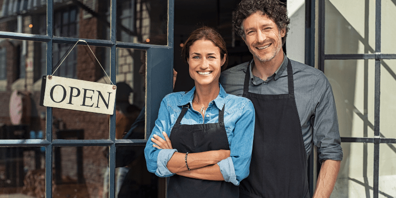 Two small business owners next to an open sign on the store door