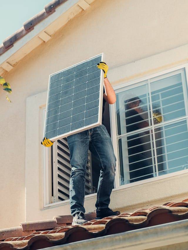 A person wearing yellow gloves holds a solar panel and stands on the sloped roof of a house next to a large window. The individual appears to be installing or preparing to install the panel, contributing to the growing solar market. The roof has a reddish-brown tile pattern.